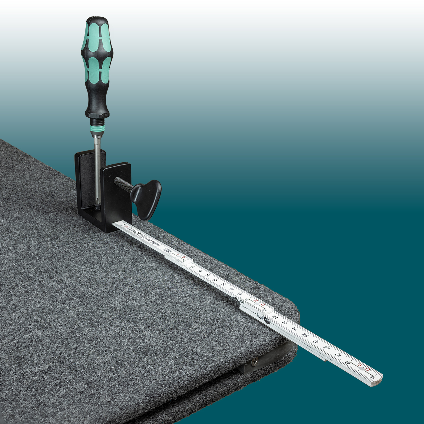 A dark grey acoustic table back panel made of felt, designed to provide sound insulation, is fitted to a table top. A black clamp secures the back panel using a green and black screwdriver. A folding rule is used to measure its length.