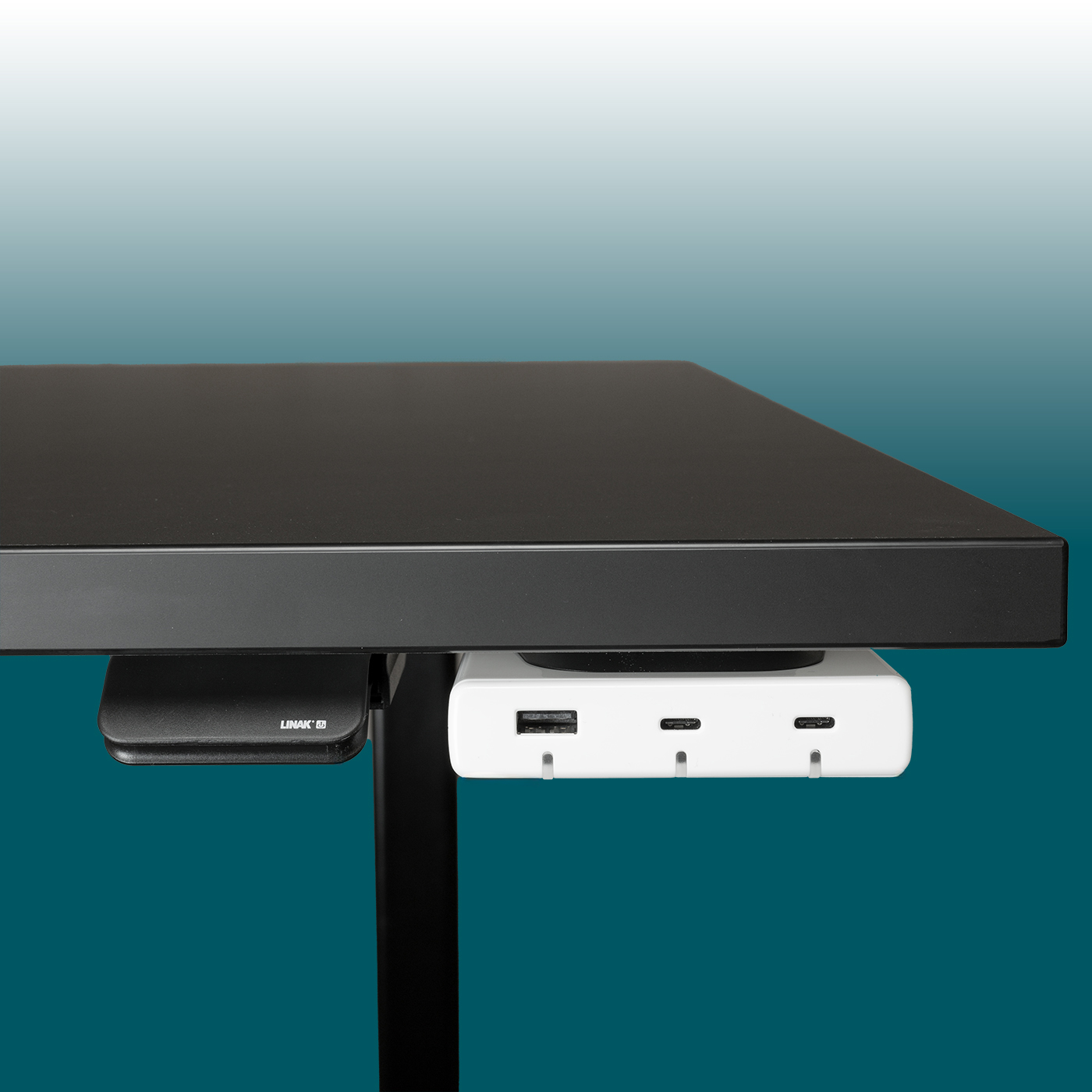 A white Desk2Device USB charger is mounted beneath a black desk top. It has three ports at the front: 1x USB-A and 2x USB-C. A black LINAK control unit, which supplies power, is attached next to it.