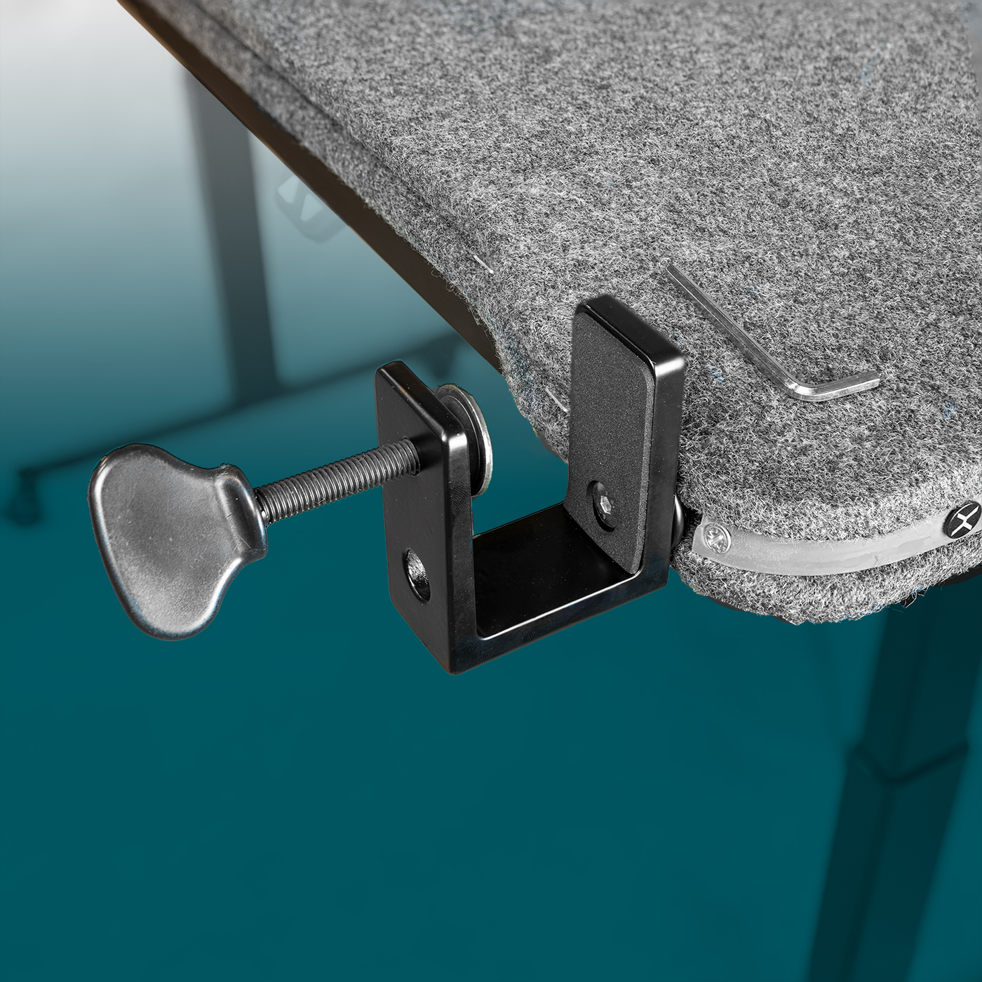 Close-up of a dark grey felt sound-absorbing table back panel, secured to the edge of the table with a black screw clamp. The clamp has a knurled wing nut. Light-coloured rubber profile with fixing points. An Allen key is provided.